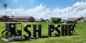 Large black metal letters spelling 'SHIPSHE' sit on a grassy field, with a vintage wagon wheel structure on the left and red barn buildings in the background under a blue sky.