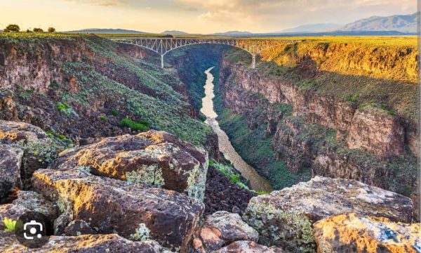 Arch bridge spanning a deep canyon with a winding river below; sunlit rocky foreground.