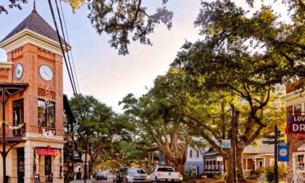 Sunlit historic main street with a red brick clock-tower building on the left, trees lining the street, and parked cars.