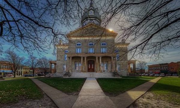 Front view of a stone town hall with a central clock tower, steps, and a grassy lawn.
