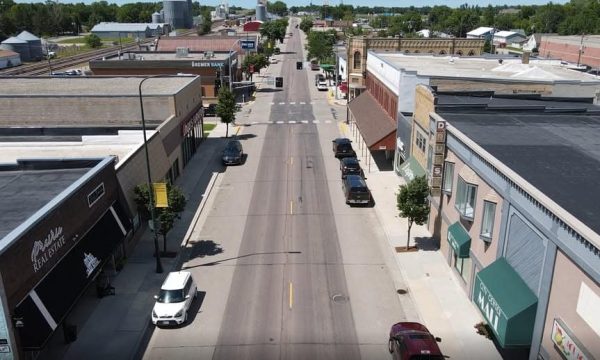 Aerial view of a small-town main street lined with storefronts, parked cars, and trees along the sidewalks.