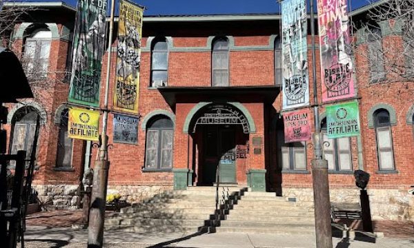 Front of a red brick museum with arched windows, steps to the entrance, and tall colorful banners on poles.